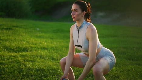 A-sporty-woman-in-a-Park-squats-with-dumbbells-in-her-hands-at-sunset.-Weight-training-in-the-open-air.-Fitness-in-a-public-Park-in-summer-for-thighs-and-legs.